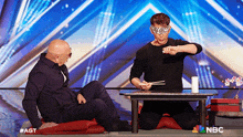 two men sitting at a table with a nbc logo on the bottom right