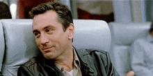 a man in a leather jacket is smiling while sitting in a chair on a plane .
