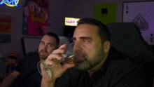 a man with a beard is drinking from a glass while another man watches .