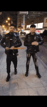 two men wearing leather jackets are standing on a sidewalk at night .