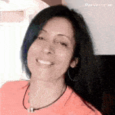 a woman with long black hair is smiling and wearing an orange shirt and necklace