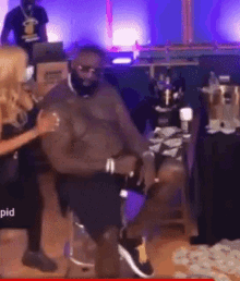 a shirtless man is sitting in a chair with a woman standing next to him