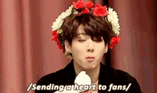 a young man wearing a flower crown on his head is holding a flower and saying `` sending a heart to fans '' .