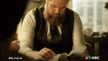 a man with a beard is reading a book with a nbc logo in the corner