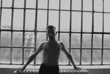 a shirtless man leans on a radiator in front of a large window