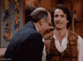 two men are standing next to each other and laughing . one of the men is wearing a vest .