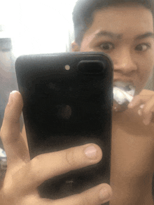 a shirtless man is brushing his teeth while taking a selfie with his iphone