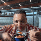 a man in a red and white shirt is eating a bowl of soup with a spoon