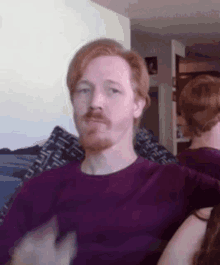 a man with red hair and a beard wearing a purple shirt is sitting in front of a mirror .