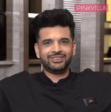 a man with a beard wearing a black shirt with a pinkvilla logo in the background