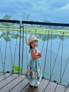 a little girl wearing a floral dress and straw hat stands on a deck overlooking a body of water