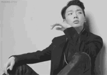 a black and white photo of a young man in a suit sitting down against a wall .
