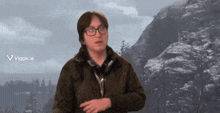a man wearing glasses and a brown jacket stands in front of a mountain