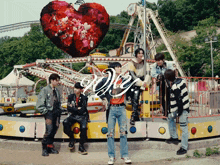 a group of young men are standing in front of a roller coaster with a heart shaped balloon in the background that says we