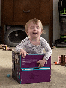 a baby is sitting in a cardboard box with a vacuum cleaner behind him