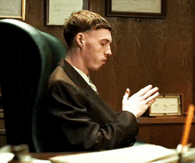a man in a suit sits at a desk with his hands folded in prayer