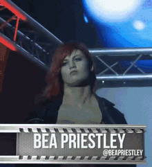 a woman with red hair is behind a sign which says bea priestley