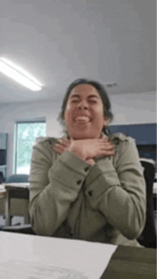 a woman in a green jacket is sitting at a desk with her hands on her chest and making a funny face .