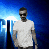 a man wearing sunglasses and a white t-shirt stands in front of a blue background with the letter c in the middle