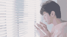 a young man in a pink shirt is looking out of a window with blinds .