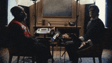 a man sits in a chair talking to a monkey who is wearing a red vest that says ' monkey '