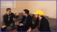 three men sitting on a couch with one wearing a yellow hat