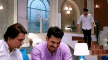 a man in a purple shirt sits in a living room with two other men