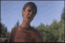 a young boy in a red shirt is standing in front of a forest with trees in the background .