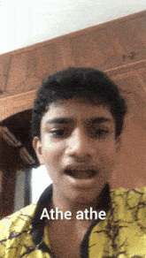 a young man wearing a yellow shirt says ' athe athe ' in front of a wooden wall