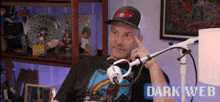 a man wearing a hat is sitting in front of a microphone with the words dark web behind him .