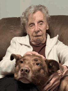 an elderly woman sits on a couch with a brown dog on her lap