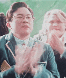 a man in a suit and glasses is clapping his hands while sitting in a crowd of people .