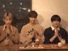 three young men are sitting at a table with their hands folded and one has a sweater that says bat in