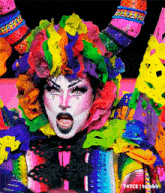 a drag queen is wearing a colorful costume with flowers on her head and horns .