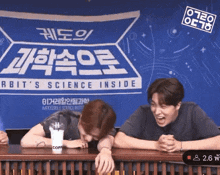 two men sitting at a table with a cup of coffee in front of a sign that says rbit 's science inside