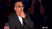 a bald man wearing sunglasses and a suit stands in front of a microphone with his hand up in the air