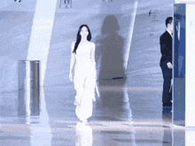 a woman in a white dress walks down a hallway with a man in a suit behind her