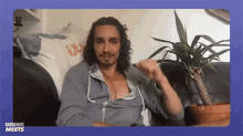 a man with curly hair and a beard is sitting on a couch with a potted plant in the background .