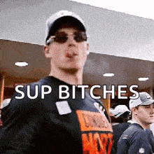 a man wearing sunglasses and a hat is smoking a cigarette and says " sup bitches " .