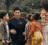 a man in a superman costume is hugging a group of children in a park .