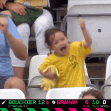 a little girl in a yellow shirt is sitting in a stadium watching a game between graham and bouchier