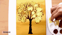 a person is painting a tree with yellow leaves on a yellow background with drawbook below it
