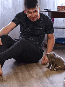 a man in a black adidas shirt sits on the floor with a puppy
