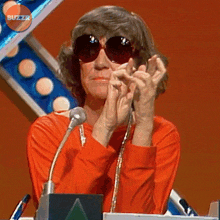 a woman wearing sunglasses is clapping her hands in front of a microphone with the buzzr logo behind her