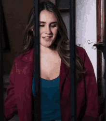 a woman in a burgundy jacket and a blue tank top smiles behind bars