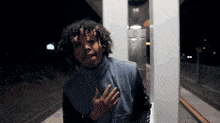 a man with curly hair is standing in front of a telephone booth with his hand on his chest