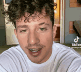 a man with curly hair and a beard is wearing a white t-shirt and smiling .