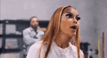 a woman with long hair and makeup is standing in a room with a man in the background .