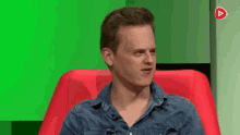 a man in a denim shirt is sitting in a red chair with a green background .