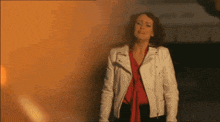 a woman in a white leather jacket is standing in front of a burning building
